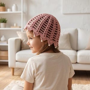 May include: A dusty rose crochet bonnet featuring a vertical ribbed pattern and a scalloped edge adorned with small white beads along the trim. The headwear is crafted with an openwork design, providing a textured appearance with delicate bead accents.