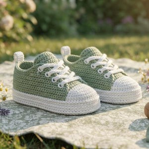 May include: A pair of sage green crochet baby sneakers featuring white laces, white toe caps, and white soles. The shoes have a small white pull tab at the heel and are constructed with a textured yarn stitch pattern, mimicking the look of classic lace-up athletic footwear.