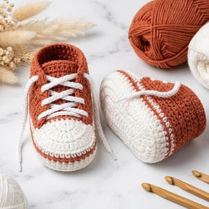 May include: A pair of handmade crochet baby booties designed to resemble sneakers, featuring a rust-orange upper and a white toe cap with white laces. The booties are accompanied by three balls of yarn in rust and white, and three wooden crochet hooks.