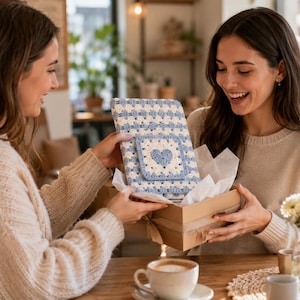 Puede incluir: Una funda rectangular de ganchillo con un patrón de cuadrados granny en azul claro y blanco, con un parche cuadrado central que muestra un motivo de corazón en azul oscuro, dentro de una caja de regalo de cartón con papel de seda.