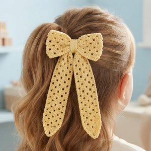 May include: A pale yellow crochet hair bow featuring a classic bow shape with two long, dangling tails. The accessory is crafted with a visible open-stitch pattern and is secured to the hair, showcasing a light, textured yarn construction.