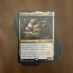 May include: A Magic: The Gathering trading card titled Kudo, King Among Bears, featuring a brown bear with antlers in a snowy forest. The card text reads: Other creatures have base power and toughness 2/2 and are Bears in addition to their other types.