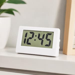 May include: A rectangular digital clock with a white plastic frame and a light green screen. The display shows the time in large, bold black digital numerals reading 12:45. The clock has a clean, simple design with a flat base for stability.