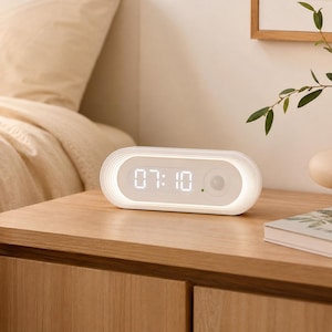 May include: A white oval-shaped digital alarm clock with a smooth, rounded frame. The front display features a bright white digital readout showing the time 07:10, a small green indicator light, and a circular sensor on the right side.