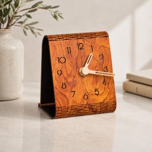 May include: A tabletop clock with a curved, wood-grain face featuring laser-cut black numerals from one to twelve. The clock has two gold-toned metal hands centered on the face, and the structure is supported by a black base.