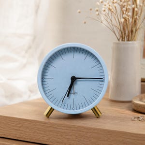 May include: A round, light blue analog clock with a minimalist face featuring black tick marks for hours and minutes. The clock has thin black hands, a quartz movement, and is supported by two small, cylindrical gold-toned metal legs.