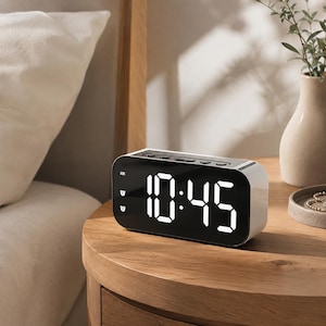 May include: A rectangular digital alarm clock with a white casing and a black front display panel. The screen shows the time 10:45 in bright white LED digits, with small icons for AM and two separate alarm settings visible on the left side.