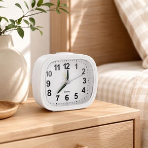 Puede incluir: Un despertador de cuarzo rectangular blanco con esquinas redondeadas, que presenta una esfera blanca, números arábigos negros, manecillas de hora y minutos negras finas y un segundero amarillo. El reloj tiene un diseño simple y limpio.