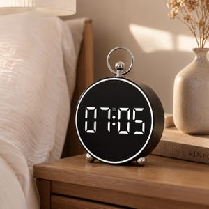 May include: A round digital alarm clock with a black face and a white LED display showing the time 07:05. The clock features a silver-toned circular handle on top, a thin silver rim around the display, and two small silver spherical feet at the base.