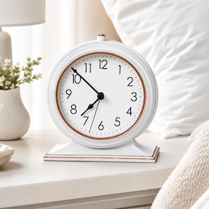 Puede incluir: Un reloj de mesa analógico blanco con esfera redonda, números negros y manecillas negras. El reloj presenta un borde interior de color cobre y una base rectangular con un borde de color cobre a juego. Un pequeño pomo de color cobre está arriba.