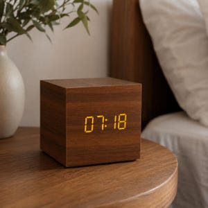 Può includere: Una sveglia digitale a forma di cubo con finitura in venature del legno marrone scuro. La parte anteriore mostra l'ora 07:18 in numeri digitali giallo brillante. L'orologio ha una struttura solida e liscia con un design squadrato e bordi netti.