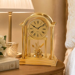 Peut inclure: Une horloge de cheminée dorée avec une finition polie, dotée d'un cadran circulaire avec des chiffres romains et des aiguilles noirs. L'horloge est soutenue par deux colonnes verticales et un sommet arqué, avec un pendule rotatif décoratif.