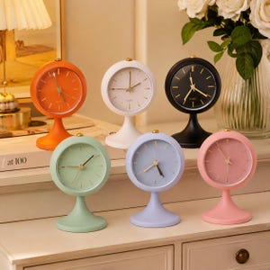 Peut inclure: Six horloges analogiques rondes sur socle disposées sur deux rangées. Les horloges sont orange, blanches, noires, vert clair, bleu clair et roses. Chaque cadran présente des index et des aiguilles dorés fins, avec le mot QUARTZ inscrit au centre.