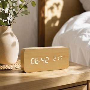 Könnte beinhalten: Ein rechteckiger digitaler Wecker mit einer hellen Holzmaserung. Das Frontdisplay zeigt leuchtend weiße LED-Zahlen, die die Uhrzeit 06:42 und die Temperatur von 21°C anzeigen. Die Uhr hat abgerundete Kanten und eine glatte Oberfläche.