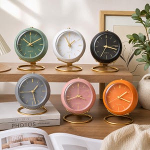 Peut inclure: Six horloges de table à quartz rondes avec des aiguilles dorées et des supports circulaires en fil d'or. Les horloges sont vert sauge, blanches, noires, grises, rose poudré et orange, avec un cadran épuré et le mot QUARTZ inscrit.