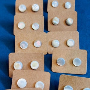 May include: A collection of round, iridescent mother-of-pearl stud earrings, each pair mounted on a small rectangular brown kraft paper card. The earrings feature a smooth, polished surface with subtle white and pearlescent color variations.