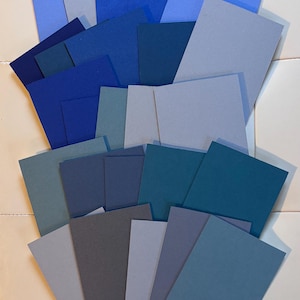 May include: A collection of rectangular cardstock sheets in various shades of blue, ranging from light sky blue and slate gray to deep navy and teal. The paper pieces are layered and overlapping, displaying a matte texture and solid color finish.