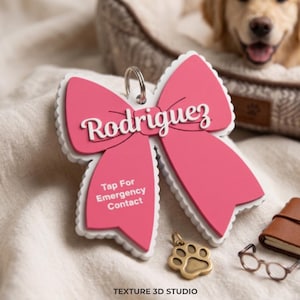 May include: A pink bow-shaped pet ID tag with a white scalloped edge, featuring the name Rodriguez in white script and the text Tap For Emergency Contact. A silver metal ring is attached to the top, and a small brass paw print charm sits nearby.