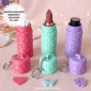 Könnte beinhalten: Drei zylindrische Lippenstifthüllen mit Schlüsselanhängern, verziert mit geprägten Herz-, Schmetterlings- und Blumenmustern in Pink, Mintgrün und Lavendel. Der Text lautet LIPSTICK CASE WITH ORNAMENT KEYCHAIN STL-3MF und TEXTURE 3D STUDIO.