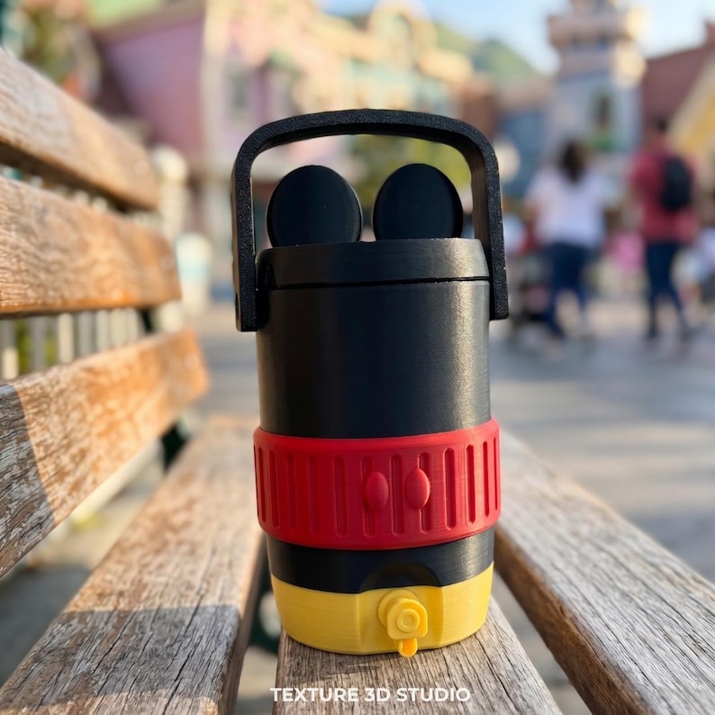 May include: A black cylindrical container with a top handle and two circular ears. It features a wide red band around the center with two vertical oval accents and a yellow base with a small protruding spout. The text TEXTURE 3D STUDIO is visible.