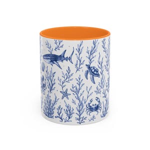 Puede incluir: Una taza de cerámica blanca con un interior naranja brillante. El exterior presenta una ilustración repetitiva en estilo de tinta azul de vida marina, incluyendo un tiburón ballena, tortuga marina, cangrejo, estrella de mar y algas.