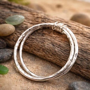 May include: A silver bangle bracelet featuring a hammered, textured surface. The design consists of two overlapping circular bands that are joined together at one point by a series of tightly wound silver wire coils, creating a layered appearance.