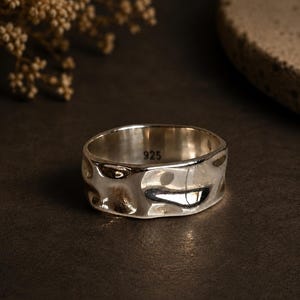 May include: A wide sterling silver ring with a polished, irregular, and hammered surface texture. The interior of the band is clearly stamped with the hallmark 925, indicating the silver content of the metal. The ring has a reflective finish.