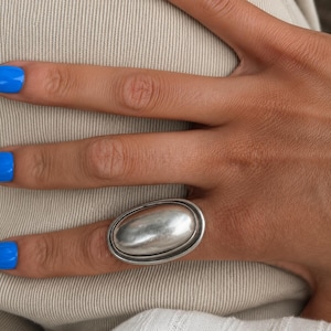May include: A large, oval-shaped silver ring featuring a smooth, polished, and slightly domed center piece framed by a thin, raised metallic border. The ring has a substantial, solid appearance with a reflective, metallic finish that catches light.