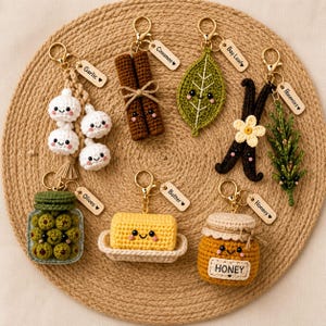 May include: A collection of seven crocheted keychains featuring food-themed designs including garlic, cinnamon sticks, a bay leaf, vanilla beans, rosemary, a jar of olives, a stick of butter, and a honey pot, each with a small labeled tag.