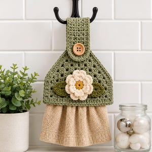 May include: A sage green crocheted towel topper featuring a central cream-colored flower with a yellow center and two dark green leaves. The topper has a wooden button closure and is attached to a beige terry cloth towel hanging from a black hook.