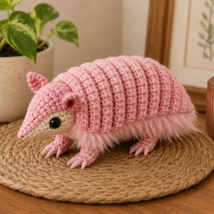 Puede incluir: Una figura de armadillo tejida a ganchillo con un caparazón segmentado de color rosa claro, un hocico color crema y ojos negros oscuros. El cuerpo está acentuado con suave piel sintética rosa claro en la parte inferior y tiene pequeñas garras.