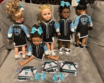 Könnte beinhalten: Fünf Puppen tragen schwarze und türkisfarbene Cheerleader-Uniformen mit weißen Streifen und Strasssteinen. Die Uniformen haben ein weißes und türkisfarbenes Logo mit den Buchstaben "NT". Die Puppen sitzen auf einem hellblauen Sofa. Es gibt drei Plastiktüten mit zusätzlichen Uniformen und Schleifen.
