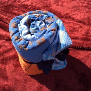 May include: A rolled up blue and brown plaid blanket with a navy blue bow. The blanket is on a red fuzzy surface.