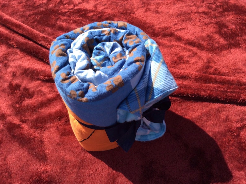 May include: A rolled up blue and brown plaid blanket with a navy blue bow. The blanket is on a red fuzzy surface.