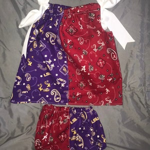 May include: A two-piece baby outfit with a purple and red bandana print. The top has a white bow and the bottom is a diaper cover. The print features the LSU and Alabama logos.