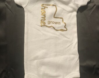 May include: White baby onesie with a gold outline of the state of Louisiana and the words "home grown" inside the outline.