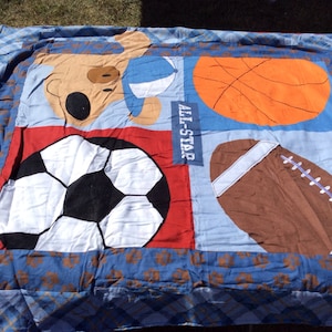 May include: A blue and red sports themed quilt with a brown bear holding a basketball, a soccer ball, a basketball, and a football. The quilt has the words "All-Star" printed on it.