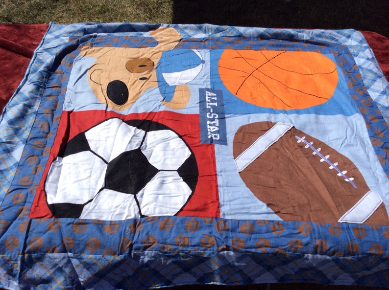 May include: A blue and red sports themed quilt with a brown bear holding a basketball, a soccer ball, a basketball, and a football. The quilt has the words "All-Star" printed on it.