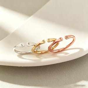 Peut inclure: Trois bagues ouvertes aux tons argent, or et or rose. Chaque bague présente une extrémité plate et effilée gravée d'une lettre majuscule. La bague argentée porte les lettres L et T, la dorée S et Z, et la bague en or rose T et H.