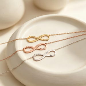 May include: Three delicate chain bracelets featuring infinity symbol pendants in gold, rose gold, and silver tones. Each pendant is engraved with two names and a date, including 08.15.2022, 03.15.2021, and 10.14.2020, displayed on a smooth, off-white circular dish.