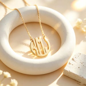 May include: A gold-toned circular pendant necklace featuring an intricate cutout monogram design with the letters R, M, and J intertwined. The pendant is suspended from a delicate gold-toned chain, showcasing a polished metallic finish.