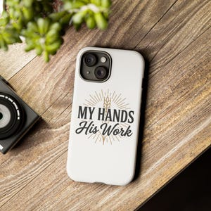 May include: A white smartphone case featuring a black graphic design with the text "MY HANDS HIS WORK" centered above a wheat stalk illustration. The design includes radiating lines in a tan color, creating a sunburst effect behind the text.