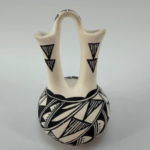 May include: A cream-colored ceramic wedding vase with two spouts connected by a central handle. The vessel features intricate black geometric patterns, including triangles on the spouts and abstract leaf-like shapes on the rounded base.