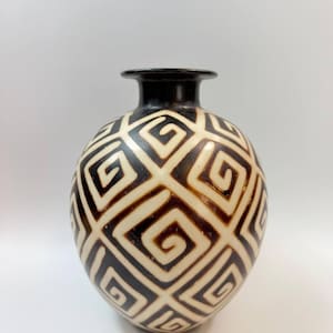 Może przedstawiać: Okrągły ceramiczny wazon z wąską szyjką i rozszerzanym brzegiem. Powierzchnia posiada powtarzający się geometryczny wzór ciemnobrązowych, zazębiających się spiralnych kwadratów na kremowym tle, tworząc efekt wizualny o wysokim kontraście.