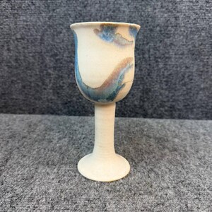 Puede incluir: Una copa de cerámica con borde acampanado y tallo delgado sobre una base circular. El exterior presenta un acabado mate en color blanco roto con vetas arremolinadas de esmalte azul y marrón que envuelven el cuenco, creando un aspecto jaspeado.