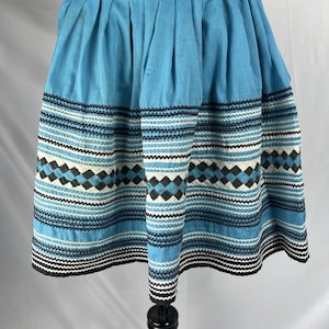 May include: A pleated light blue skirt featuring horizontal bands of black and white rickrack trim and a central row of black and white diamond-shaped patterns. The skirt has a solid light blue waistband and a hem finished with black rickrack.