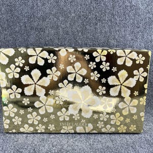 Puede incluir: Una caja rectangular de Estée Lauder con un acabado dorado metálico y un patrón repetitivo de flores blancas y amarillo claro de cinco pétalos. El nombre de la marca Estée Lauder está impreso en letras mayúsculas blancas en la parte inferior.