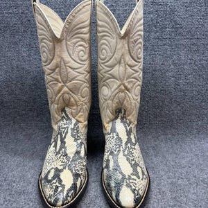 Könnte beinhalten: Ein Paar hohe Stiefel im Western-Stil mit einem cremefarbenen Lederschaft mit aufwendigen Ton-in-Ton-Nähten und einer spitz zulaufenden Zehenpartie, die mit einem strukturierten, grau-cremefarbenen Schlangenledermuster und dunklem Sohlenrand versehen ist.