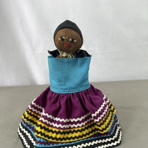 May include: A handcrafted doll with a dark brown coconut shell head, wearing a black head covering, a bright blue bodice, and a purple skirt. The skirt features horizontal rows of yellow, black, white, and light blue zigzag trim along the hem.