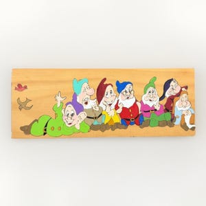 May include: A rectangular wooden plaque featuring a hand-painted illustration of seven cartoon dwarves in various colourful outfits, including green, red, blue, and purple, with two small birds flying on the left side of the light-toned wood surface.